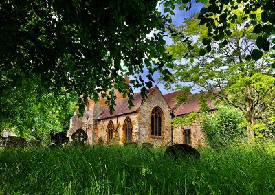 Harbury Church through the trees with headstones in the long grass, by Maureen Handle